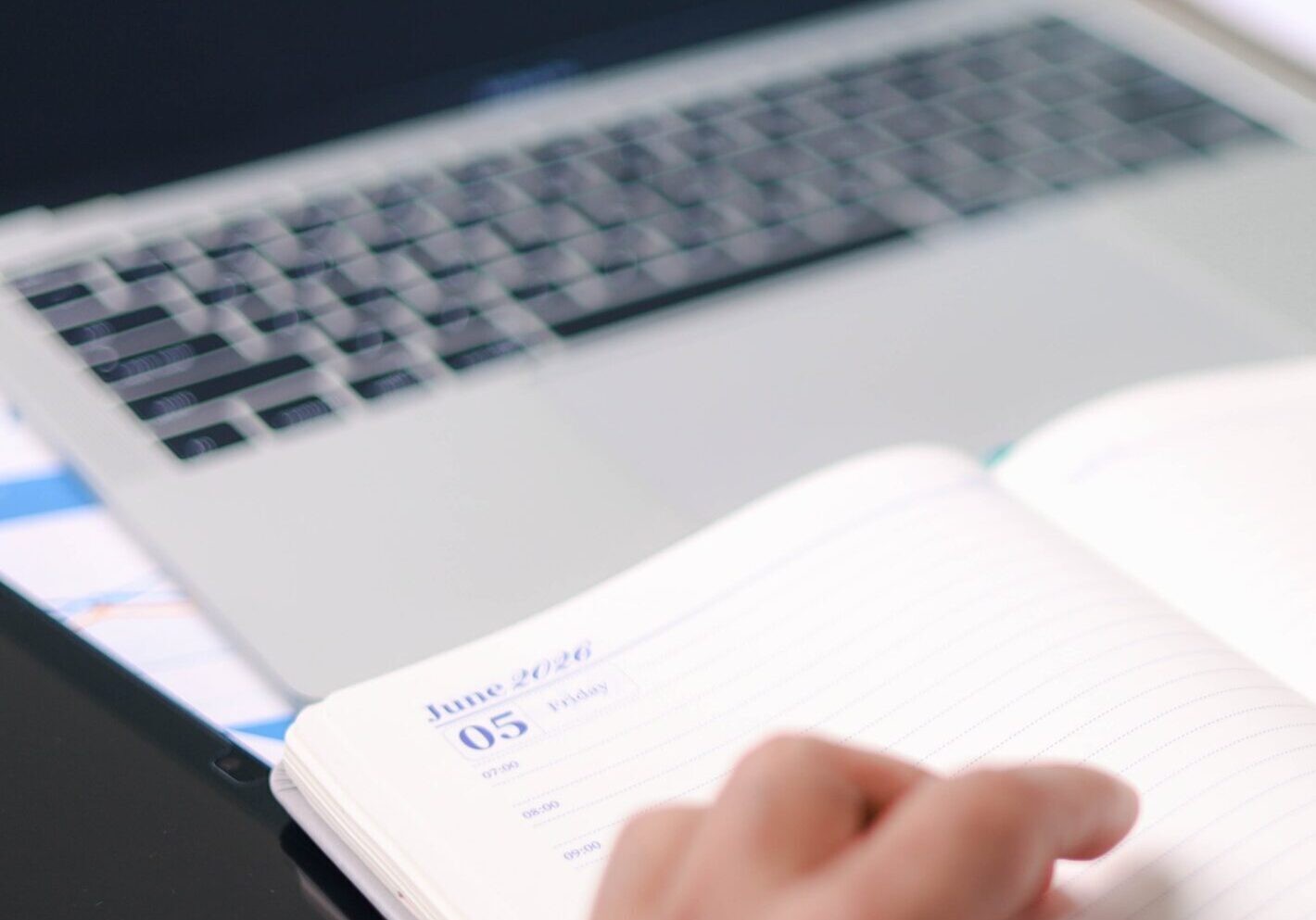 A close-up of a person's hands writing in an open planner dated June 2026 with a pink pencil, with a blurred laptop and charts visible in the background.