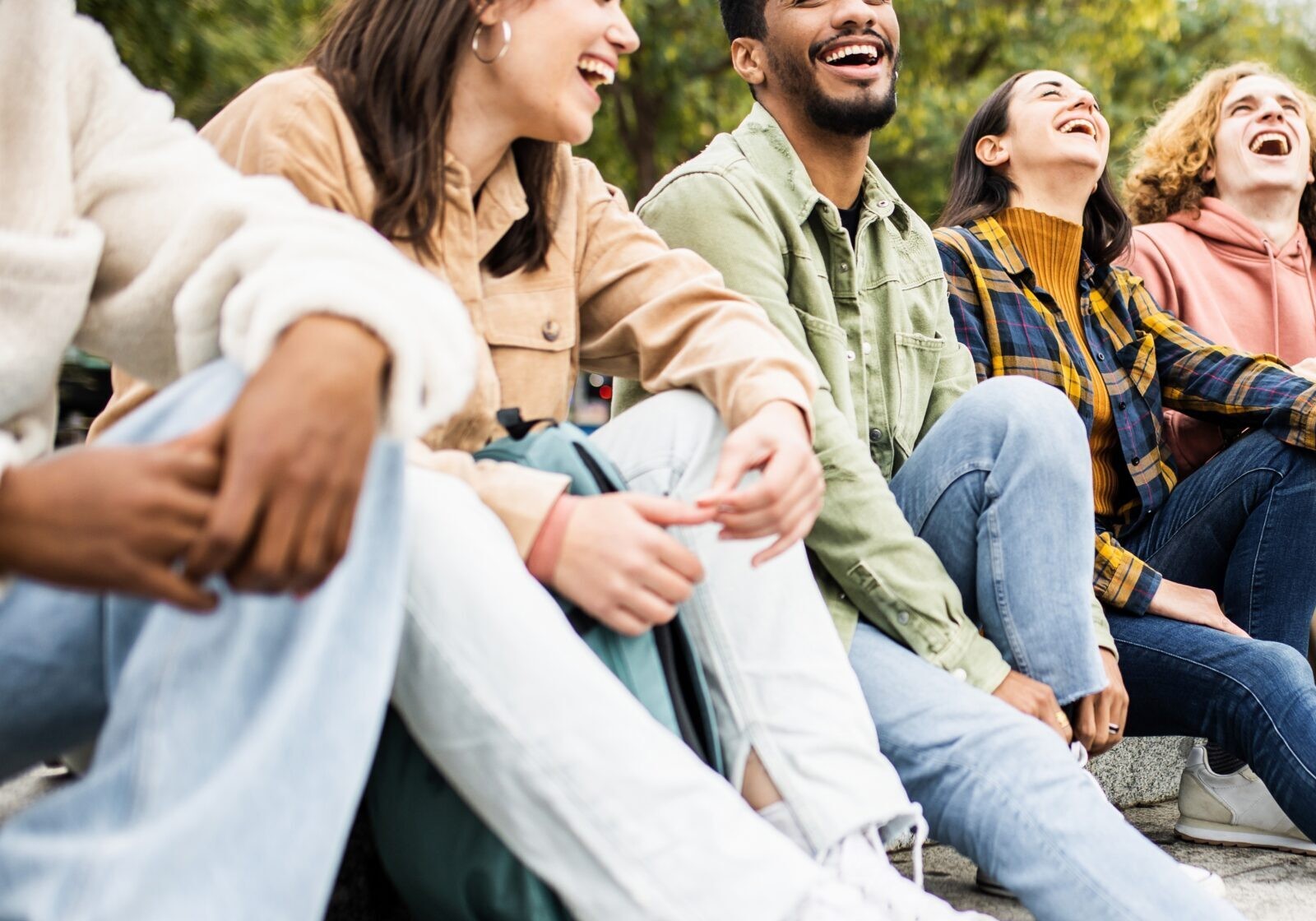 A diverse group of six young friends sitting together on outdoor stone steps, laughing and enjoying a casual conversation in a park.