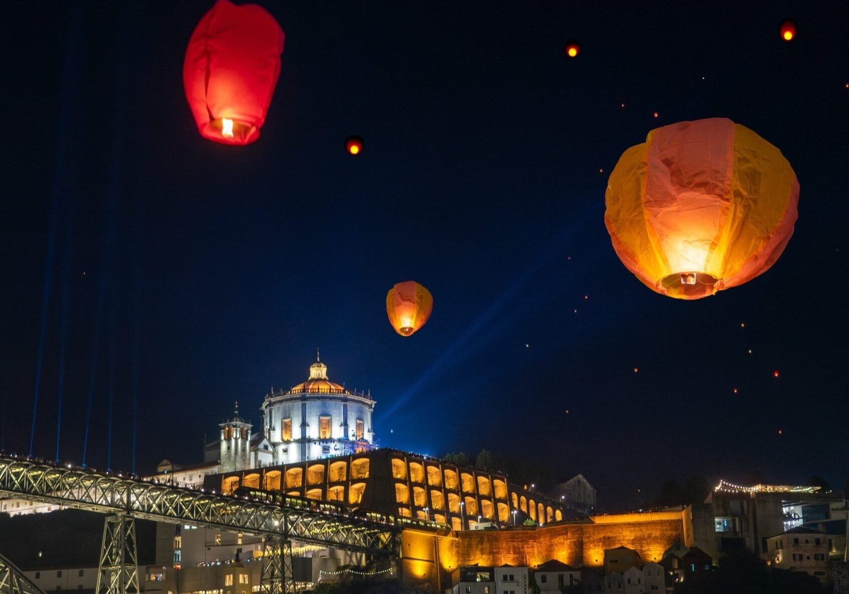 Glowing sky lanterns float in the dark night sky above a city featuring a large arched bridge and an illuminated hilltop building.