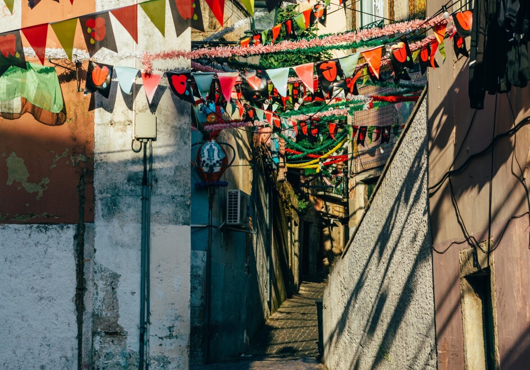 A narrow, steep, cobblestone alleyway between weathered buildings, adorned with vibrant strings of triangular bunting flags and thick red and white tinsel garlands strung overhead under strong, bright sunlight.