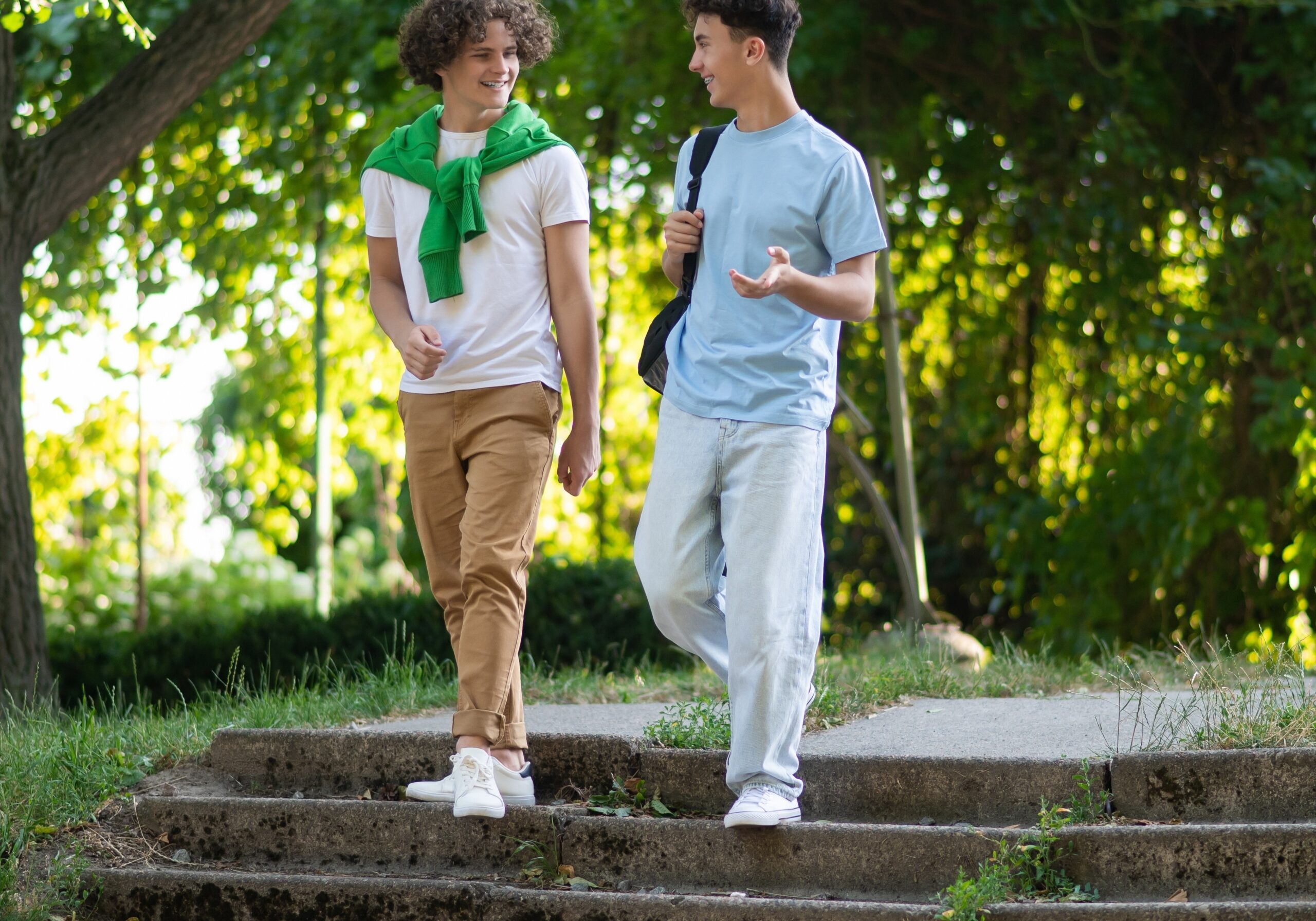 Two young men walking down a set of concrete steps outdoors, engaged in conversation and smiling at each other. One has curly hair, a white t-shirt, tan trousers, and a bright green sweater tied around his shoulders. The other wears a light blue t-shirt, loose light jeans, and a black backpack. They are surrounded by lush green trees and foliage with dappled sunlight filtering through.