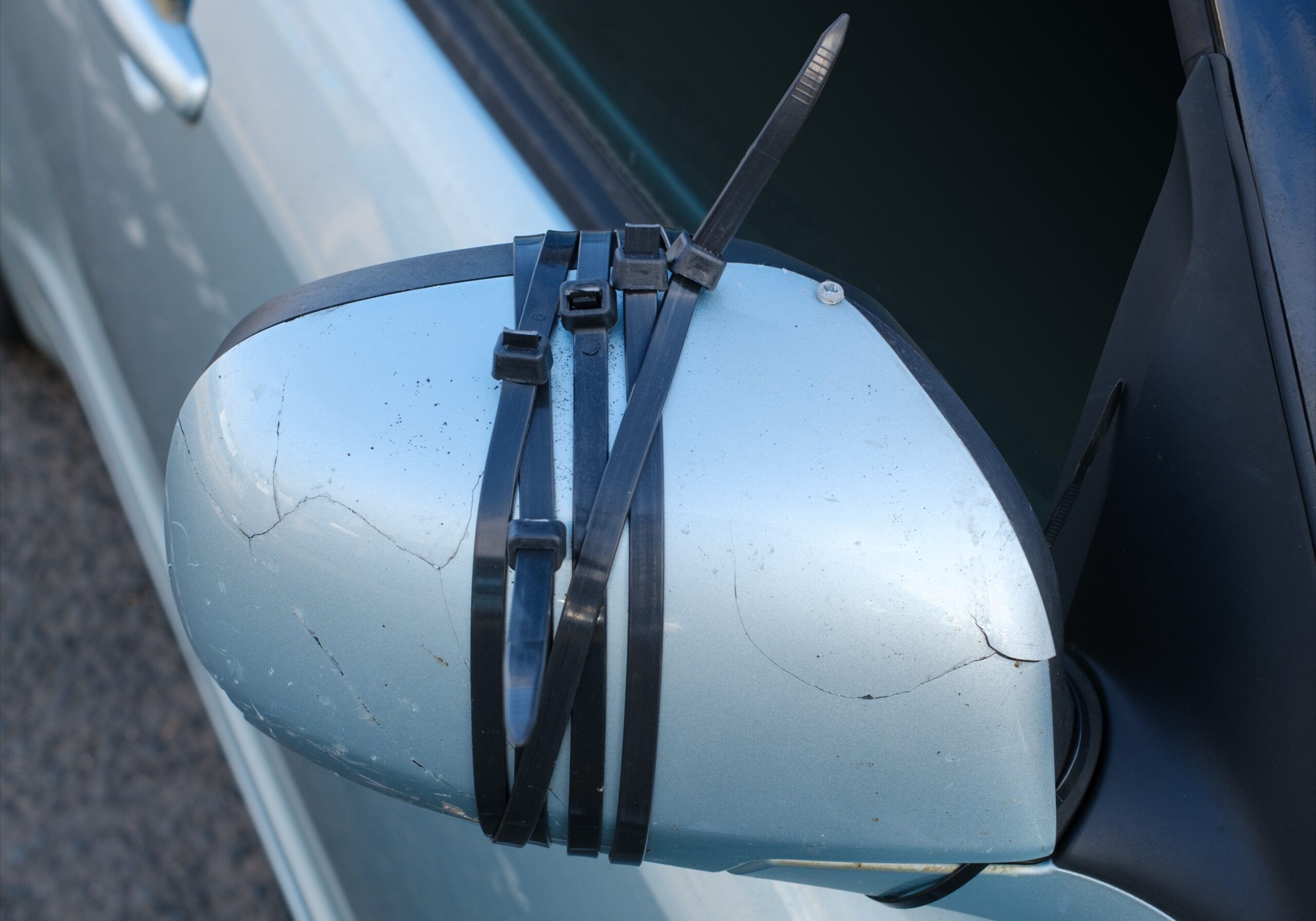 close-up of a light blue car's side-view mirror that has been damaged. Several black zip ties are wrapped tightly around the cracked plastic housing to hold it together in a makeshift repair.