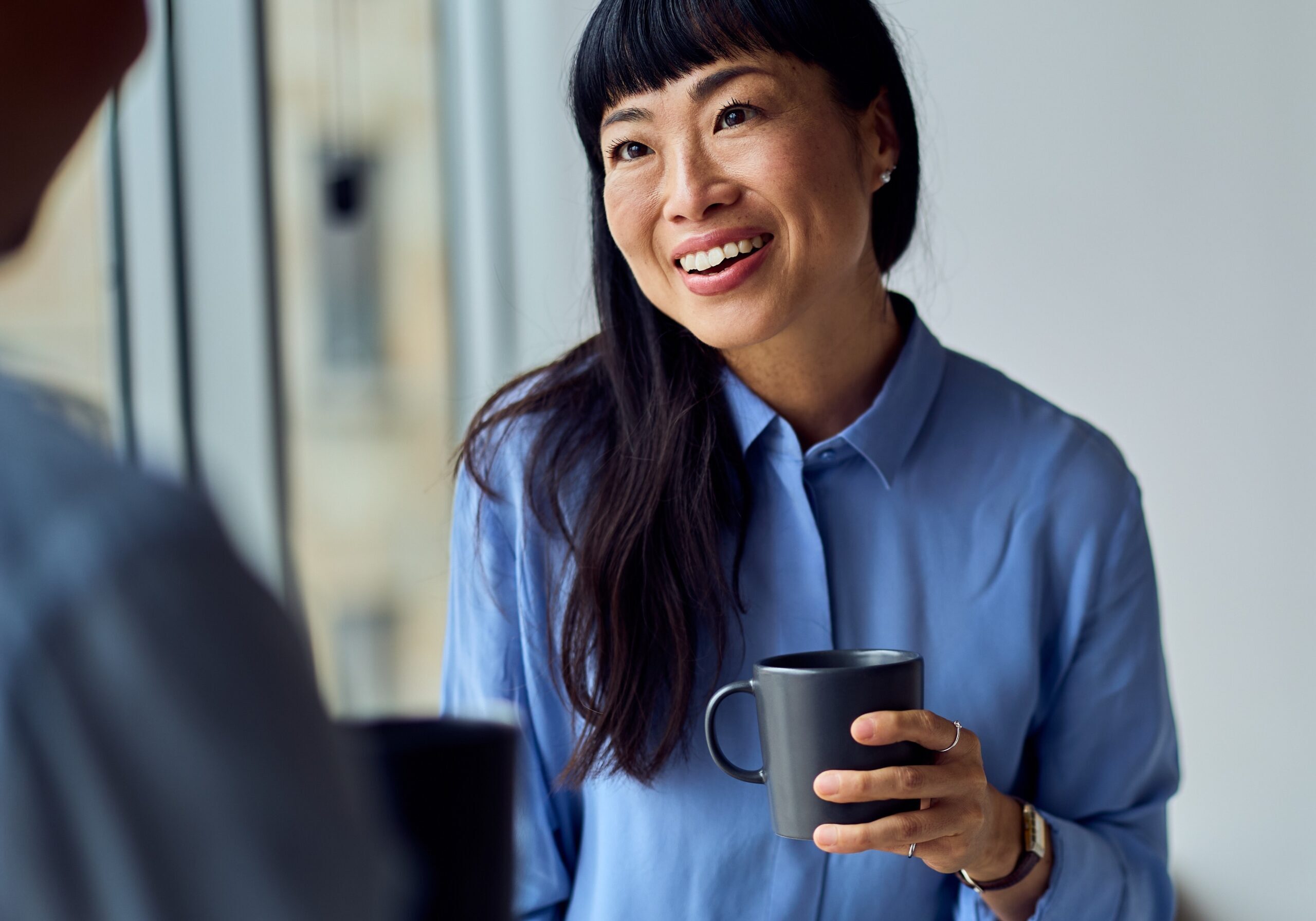 A woman with long dark hair and bangs smiles warmly while holding a grey mug. She is wearing a light blue button-up shirt and is engaged in a conversation with another person, who is partially visible in the blurred foreground