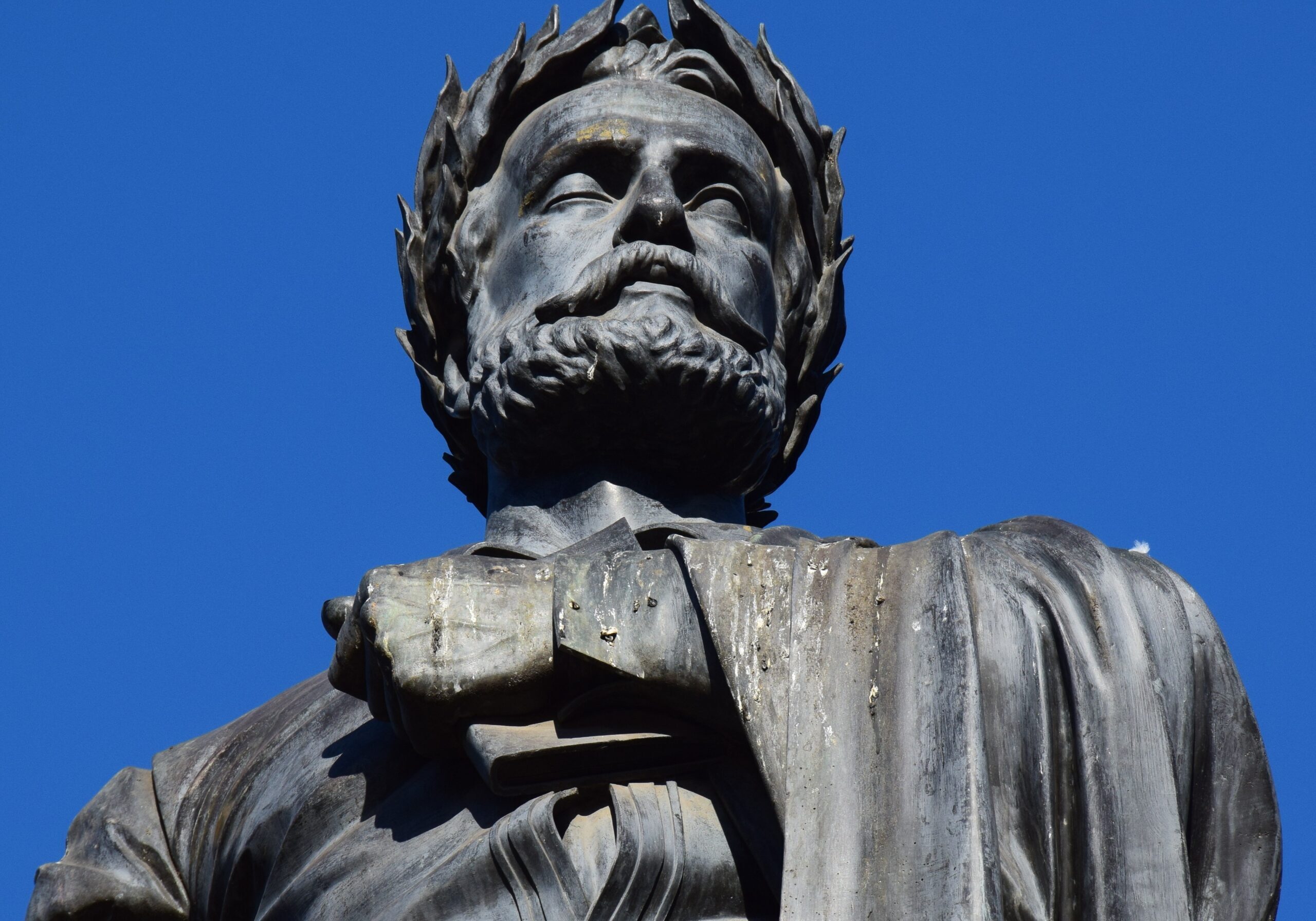 A low-angle shot looking up at a weathered bronze or dark metal statue of Camões. The statue is positioned against a clear, bright blue sky.