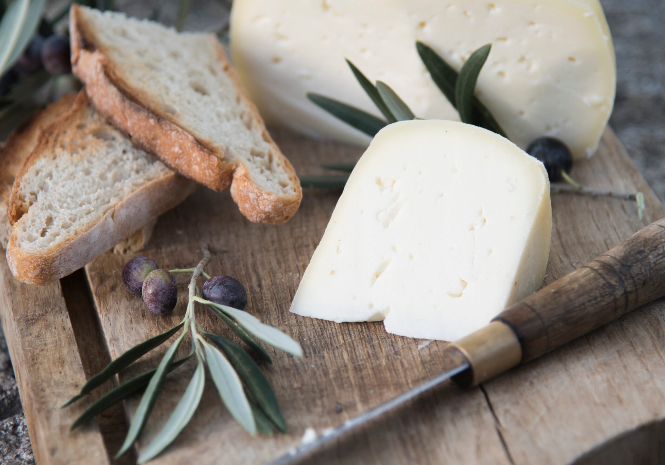 A rustic wooden board with a wheel and wedge of white cheese, toasted bread, and olive branches