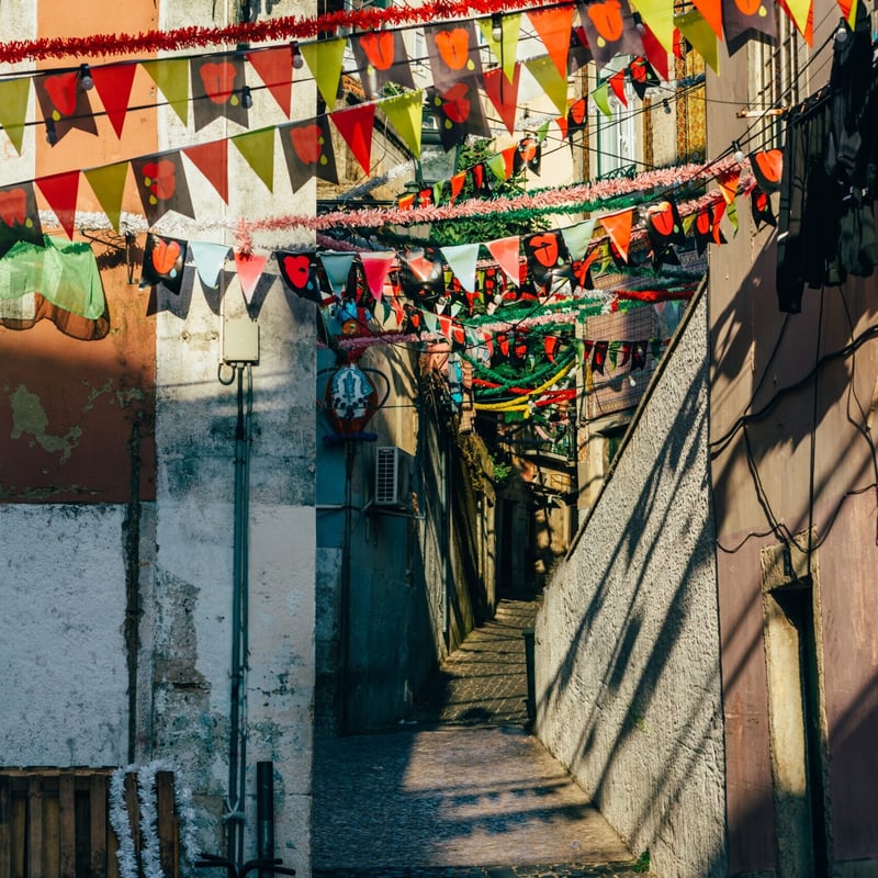A narrow, steep, cobblestone alleyway between weathered buildings, adorned with vibrant strings of triangular bunting flags and thick red and white tinsel garlands strung overhead under strong, bright sunlight.