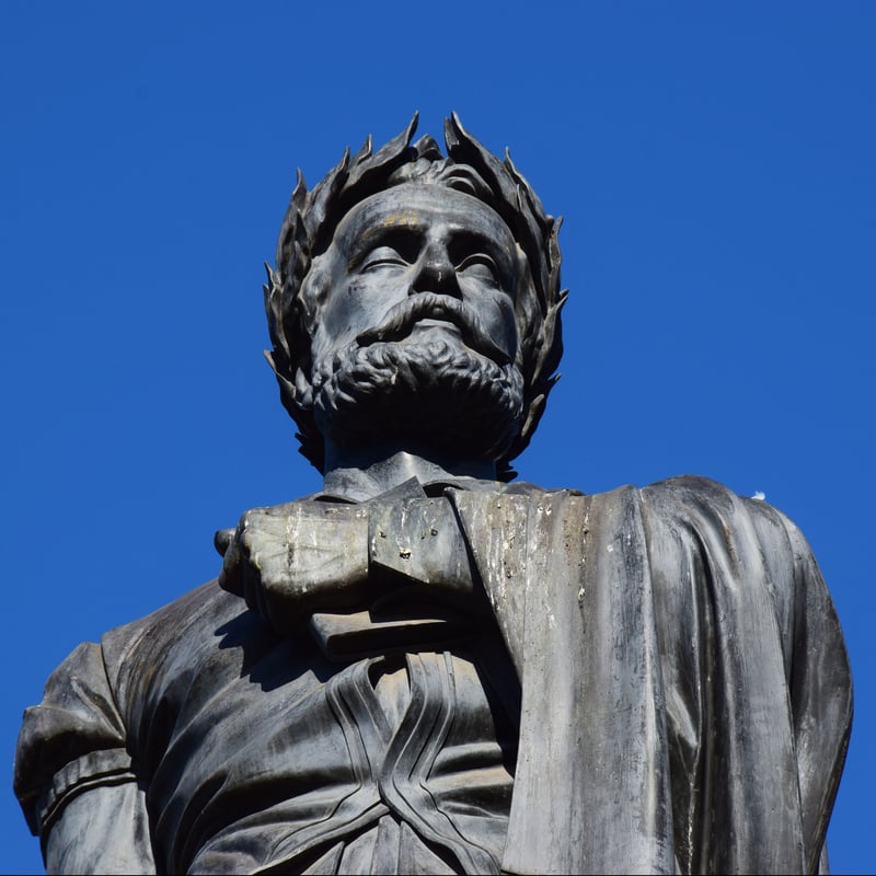 A low-angle shot looking up at a weathered bronze or dark metal statue of Camões. The statue is positioned against a clear, bright blue sky.