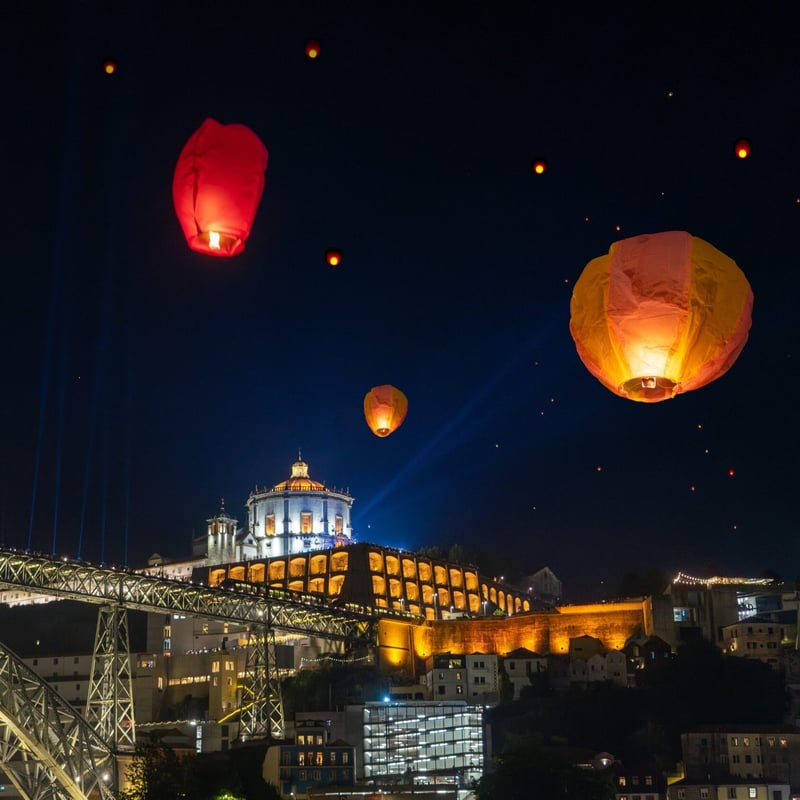 Glowing sky lanterns float in the dark night sky above a city featuring a large arched bridge and an illuminated hilltop building.