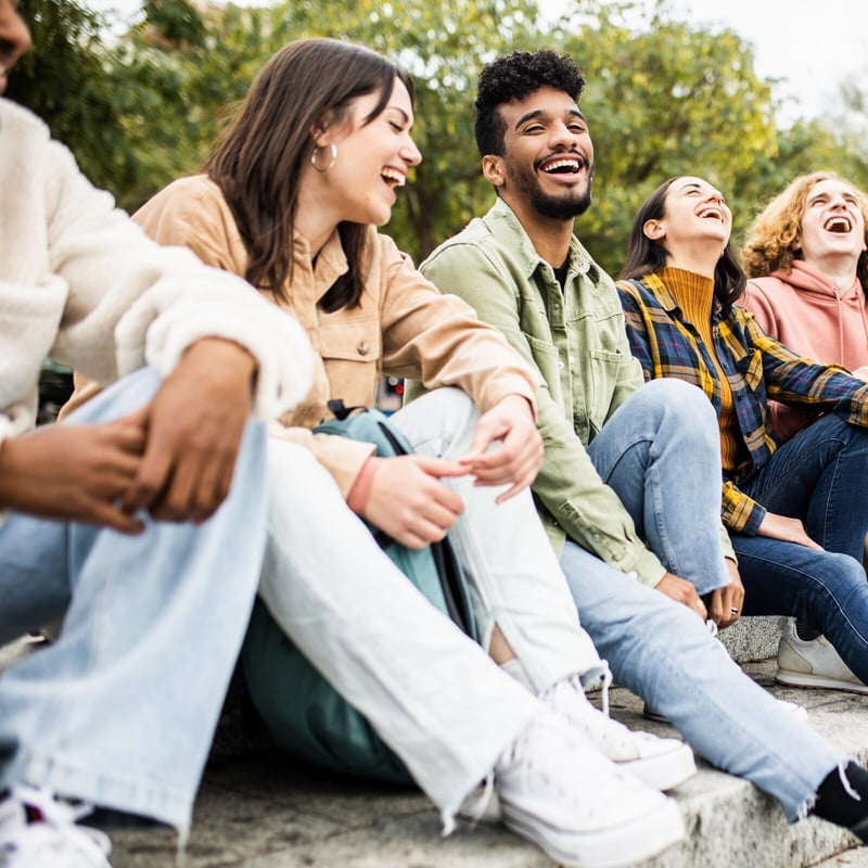 A diverse group of six young friends sitting together on outdoor stone steps, laughing and enjoying a casual conversation in a park.